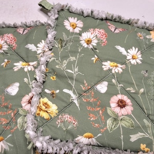 May include: Two square pot holders with a floral design featuring daisies, butterflies, and bees on a green background. The edges are trimmed with a gray, fringed fabric. Each pot holder has a green fabric loop.