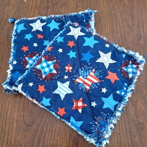 May include: Two square, patriotic-themed potholders. The navy blue fabric is decorated with red, white, and blue stars of various sizes and patterns. The edges are frayed, giving a rustic look. The potholders are likely used in the kitchen.