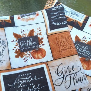 May include: Two quilted fabric squares with a fall theme. The squares feature a patchwork design with pumpkins, leaves, and phrases like "Gather Together" and "Give Thanks" in elegant script. The color palette includes orange, brown, and cream.