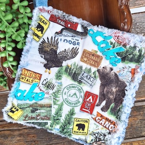 May include: A square, white and blue camping-themed decorative pillow with various patches featuring bears, eagles, and signs. The pillow has a fluffy, light blue border and the word "lake" in turquoise.