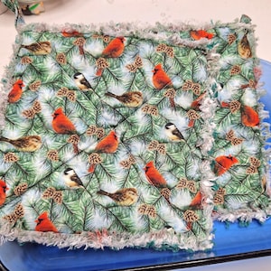 May include: A quilted potholder with a repeating pattern of red cardinals, brown birds, and small black and white birds on a background of green pine needles and pine cones. The potholder has a fringed edge.