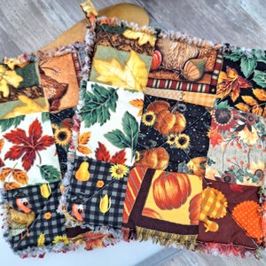 May include: A pair of square quilted potholders featuring a patchwork design with autumn-themed fabric patterns including orange pumpkins, yellow sunflowers, red maple leaves, turkeys, and black and white buffalo check squares with frayed edges.
