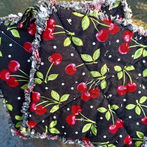 May include: Two black quilted pot holders with a cherry pattern. The pot holders feature a repeating design of red cherries, green leaves, and white polka dots. The edges are fringed with a mix of black, white, and red threads.