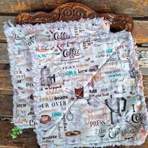 May include: Two square, quilted coffee-themed pot holders with a white background and a repeating pattern of coffee-related words and images. The edges are trimmed with a fluffy, light-colored fabric. The pot holders are resting on a decorative wooden stand.