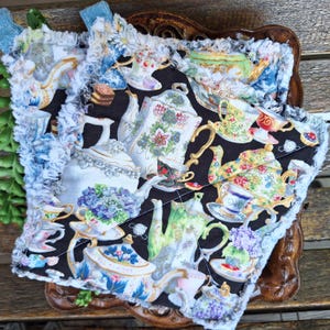 May include: A quilted potholder featuring a black background with a pattern of teacups, teapots, and floral designs in various colors. The potholder has a ruffled, light blue and white trim and hanging loops.