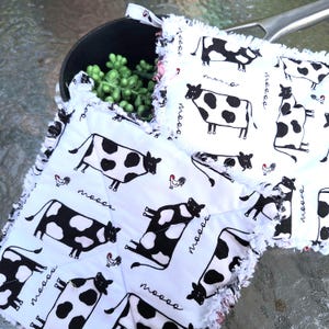 May include: White potholders with a black and white cow print and a frayed edge. The potholders have a quilted design and feature the word "moooo" and small rooster illustrations.
