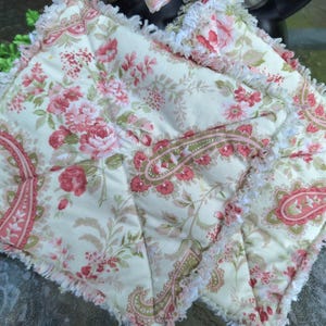 May include: Two quilted pot holders with a floral and paisley pattern in shades of pink, red, and green on a cream background. The edges are trimmed with a white fringe.