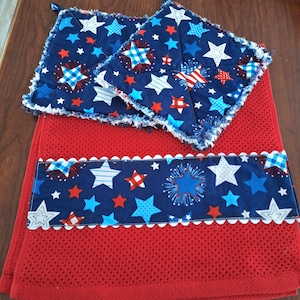 May include: A red kitchen towel and two matching pot holders. The towel has a navy blue band with white, red, and blue stars and fireworks. The pot holders and band feature a white scalloped edge.