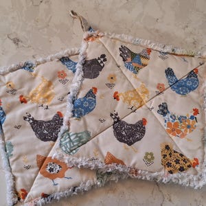 May include: Two white cotton pot holders with a frayed edge. The pot holders feature a repeating pattern of colorful chickens on a white background.