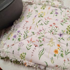 May include: A square, quilted pot holder with a white background and a floral pattern featuring various wildflowers in shades of pink, purple, yellow, and orange. The edges are trimmed with a white fringe with green accents. A dark gray pot is partially visible in the upper left corner.