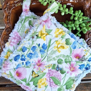 May include: Two floral print pot holders with a white background, featuring pink, yellow, and blue flowers, and butterflies. The pot holders have a ruffled edge and a loop for hanging. The pot holders are square shaped.