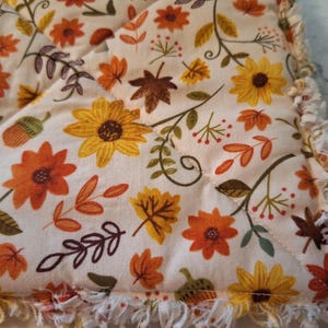 May include: A quilted fabric with a cream background, featuring a repeating pattern of sunflowers, orange and yellow flowers, acorns, and fall leaves in shades of orange, brown, and green. The edges are finished with a fringed trim.