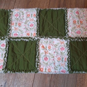 May include: A patchwork quilt runner featuring alternating green and floral print squares. The floral squares display pink roses, orange butterflies, and green foliage on a white background. The edges have a frayed, textured finish.