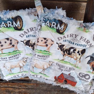 May include: Two quilted pot holders with a farm theme. Each features a blue tractor, farm animals, and text that reads "Dairy Farm Artisan Produce" and "All-Day Market". The edges are fringed.