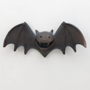 May include: A dark brown, matte-finished ceramic wall decoration shaped like a stylized bat with outstretched wings. The bat features a wide, open mouth revealing two pointed fangs, small circular eyes, and pointed ears, creating a symmetrical form.