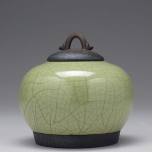 May include: A round ceramic jar with a light green crackle glaze finish. The jar features a dark brown base and a matching dark brown lid with a prominent arched handle on top. The surface of the green body displays a network of fine lines.