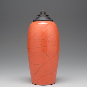 May include: A tall, orange ceramic jar with a dark gray lid and handle. The jar has a crackle glaze pattern. The lid is a dark gray color. The jar is a decorative item.