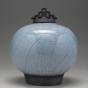 May include: A light blue ceramic jar with a crackle glaze pattern. The jar has a round body and a dark gray base. A dark gray lid with a decorative top sits on the jar. The jar is a decorative item.
