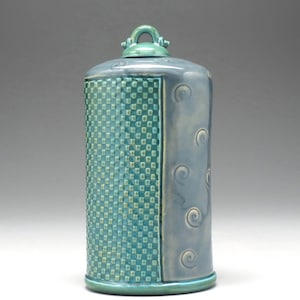 May include: A cylindrical ceramic canister with a glossy finish, featuring a teal section with a raised square grid pattern and a light blue section decorated with embossed spiral motifs, topped with a matching teal lid with a small handle.