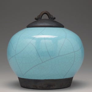May include: A round, light blue ceramic jar with a dark brown lid and base. The jar has a crackle glaze pattern. The lid features a small, arched handle. The jar is a decorative item.
