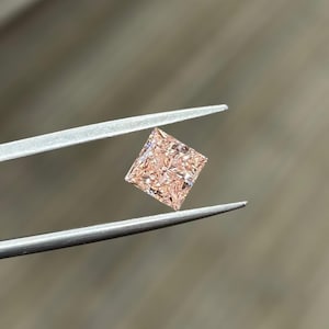 May include: A square-cut, light pink diamond held by silver tweezers. The diamond has many facets, reflecting light. The background is a blurred, neutral tone. The diamond is a gemstone.