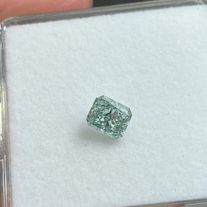 May include: A rectangular, light teal-colored diamond with a faceted cut. The gemstone is set against a white background inside a clear plastic case. The diamond's facets reflect light, creating a sparkling effect.