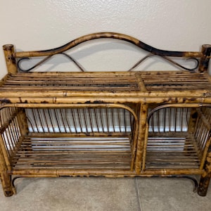 May include: A vintage bamboo bench with a natural finish. The bench features a slatted top and two open storage compartments. The back has a curved design with decorative metal accents. The bamboo has dark brown accents.