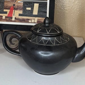 Könnte beinhalten: Eine schwarze Keramik-Teekanne mit Deckel und weißem geometrischen Design. Die Teekanne hat einen gebogenen Griff und Ausguss. Die Teekanne steht auf einer weißen Oberfläche.