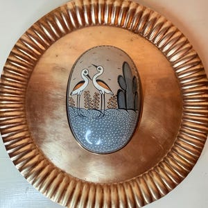 Könnte beinhalten: Ein rundes, kupferfarbenes Metalltablett mit geriffeltem Rand und einem mittig platzierten ovalen Objekt. Das ovale Stück zeigt eine bemalte Szene mit zwei langhalsigen Vögeln in einer gepunkteten Landschaft neben einem hohen Kaktus.