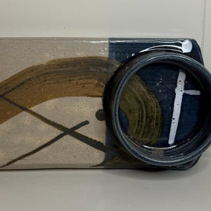 May include: A rectangular ceramic serving board with a handle, featuring a split design of beige and dark blue glaze. The surface is decorated with abstract brown brushstrokes and a white line accent, including a circular bowl integrated on the right.
