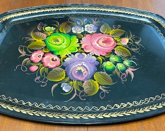May include: Oval black serving tray with a floral design. The tray features a central bouquet of pink, purple, and green flowers with green leaves. The rim is decorated with a gold-colored, repeating pattern.