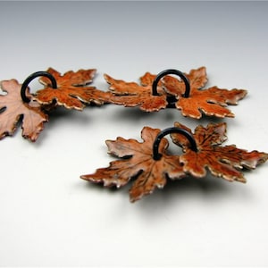 May include: Three copper maple leaf charms with black metal rings. The leaves are a deep orange color with a glossy finish.