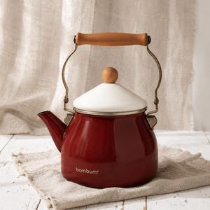 Op de afbeelding: Een rode emaille theeketel met een witte deksel en houten handvat. De ketel heeft een tuit en een metalen handvat. De merknaam "bombum" staat op de zijkant. De ketel staat op een beige doek.