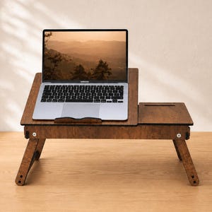 Könnte beinhalten: Ein dunkelbrauner Laptop-Schreibtisch aus Holz mit klappbaren Beinen, einer verstellbaren, abgewinkelten Plattform für einen Laptop und einem flachen Seitenteil mit rechteckigem Ausschnitt. Der Laptop-Bildschirm zeigt eine Berglandschaft bei Sonnenuntergang.