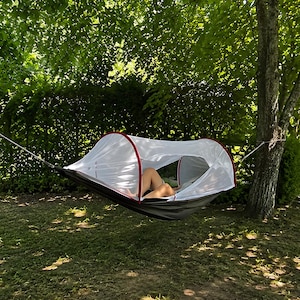 May include: A dark grey hammock featuring a white mesh canopy with red trim, suspended between two trees. The canopy has a curved frame and an open side, providing a covered space while hanging above the ground in a shaded outdoor area.