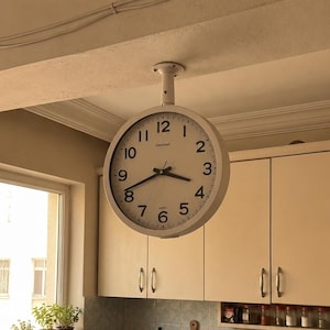 May include: A double-sided, round, white wall clock with a black frame and black hands is mounted to the ceiling. The clock face features black Arabic numerals from 1 to 12, a minute track, and the text "Westclox" and "Quartz" printed in black.