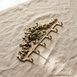 May include: A decorative brass wall hook with floral detailing. The hook has five curved hooks for hanging items. The brass finish has a vintage look. The hook is on a neutral-colored linen surface.