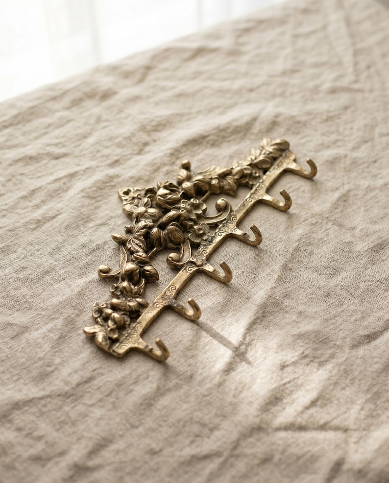 May include: A decorative brass wall hook with floral detailing. The hook has five curved hooks for hanging items. The brass finish has a vintage look. The hook is on a neutral-colored linen surface.