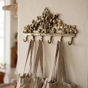 May include: Ornate brass wall hook with five curved hooks, adorned with floral and leaf details. Three beige tote bags hang from the hooks. The wall is painted a neutral color, creating a vintage aesthetic.