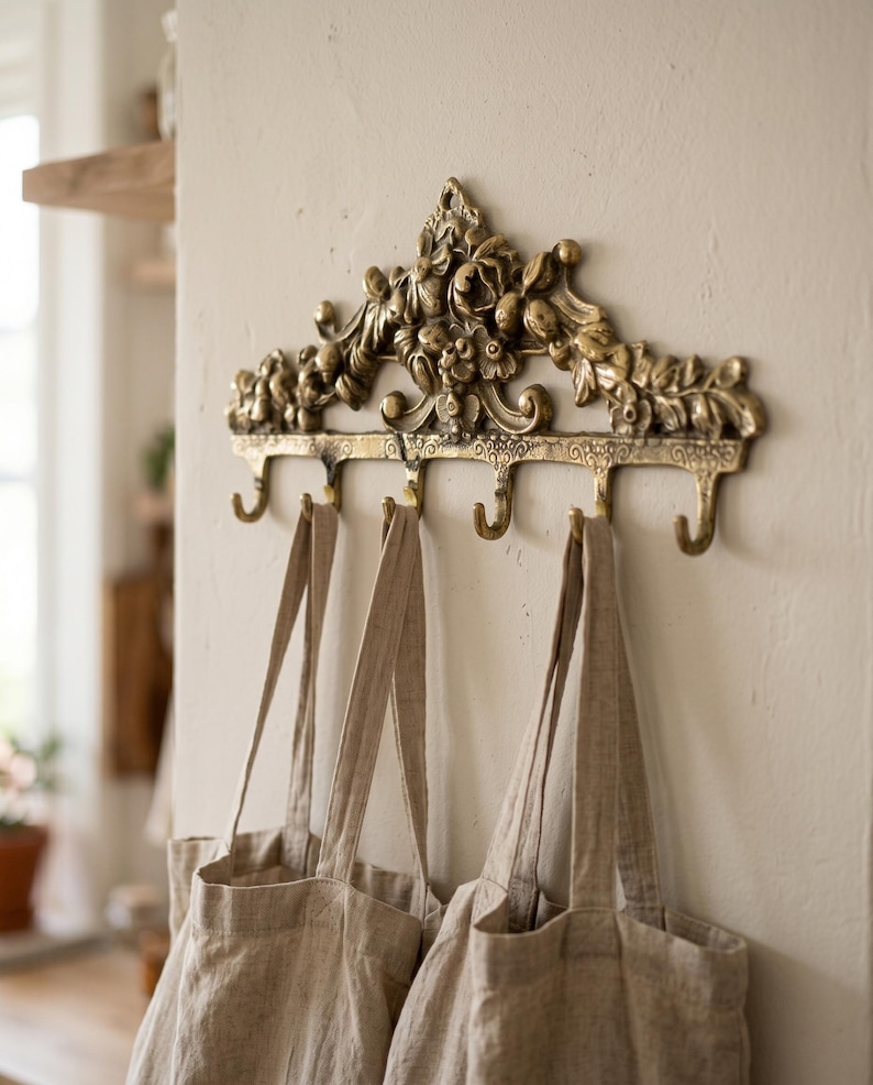 May include: Ornate brass wall hook with five curved hooks, adorned with floral and leaf details. Three beige tote bags hang from the hooks. The wall is painted a neutral color, creating a vintage aesthetic.