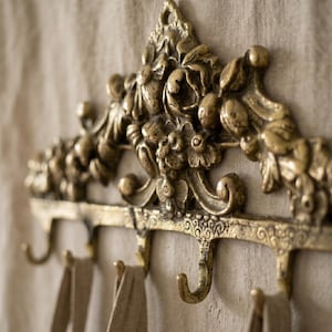May include: Ornate, antique-style wall-mounted coat rack in a warm brass finish. The rack features decorative floral and scrollwork details, with multiple curved hooks for hanging items. The rack is mounted on a neutral-colored fabric background.