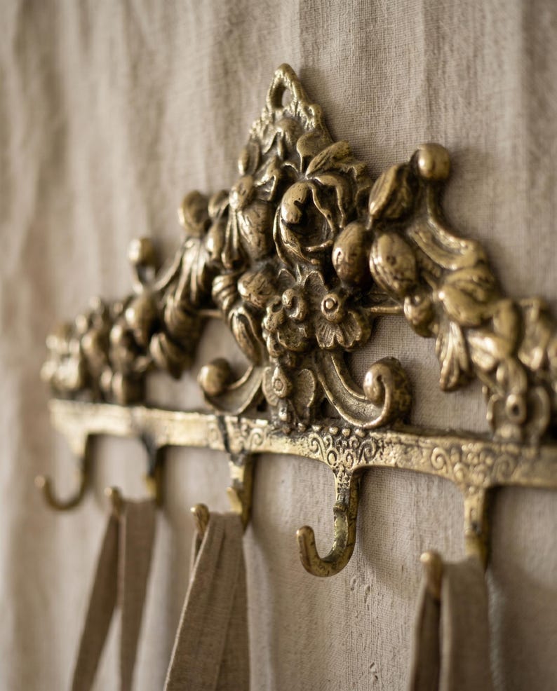 May include: Ornate, antique-style wall-mounted coat rack in a warm brass finish. The rack features decorative floral and scrollwork details, with multiple curved hooks for hanging items. The rack is mounted on a neutral-colored fabric background.