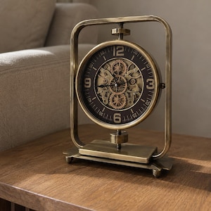 May include: A brass-toned metal table clock featuring a rectangular frame and a circular face. The clock face has a dark background with gold-coloured numerals at 12, 3, 6, and 9, and a visible internal gear mechanism in the centre with black hands.