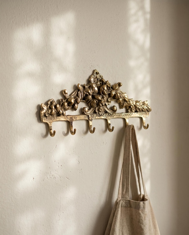 May include: A gold-colored wall-mounted coat rack with floral and leaf detailing. The rack has five hooks and a beige tote bag hanging from one of them. The rack is mounted on a light-colored wall.