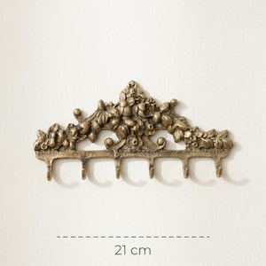 May include: A bronze-colored wall-mounted hook rack with floral and leaf detailing. The rack has six hooks and measures 21 cm (8.3 inches) in length. The design features an ornate, vintage style, suitable for hanging items.