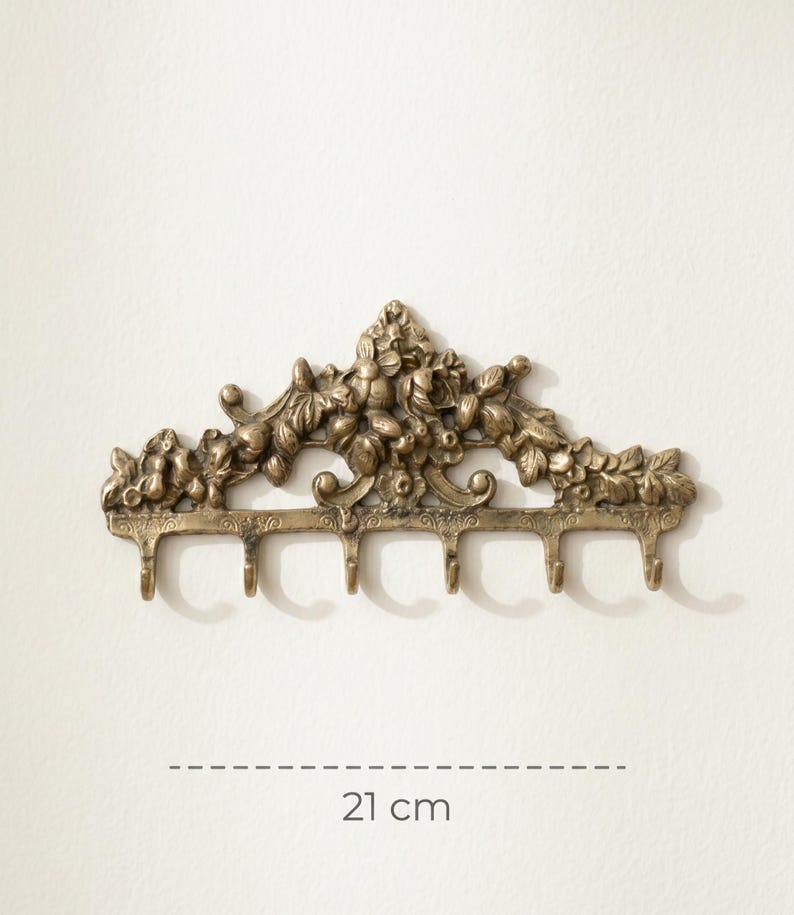 May include: A bronze-colored wall-mounted hook rack with floral and leaf detailing. The rack has six hooks and measures 21 cm (8.3 inches) in length. The design features an ornate, vintage style, suitable for hanging items.