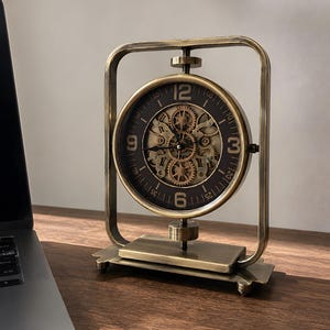 May include: A brass-toned metal table clock with a rectangular frame and a circular face. The clock features a dark brown dial with visible internal gears, black hands, and large Arabic numerals at the 12, 3, 6, and 9 o'clock positions.
