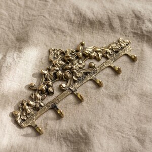 May include: A decorative brass wall hook with floral and leaf detailing. The hook has five individual hooks for hanging items. The brass has an aged appearance. The wall hook is designed to be mounted on a wall.