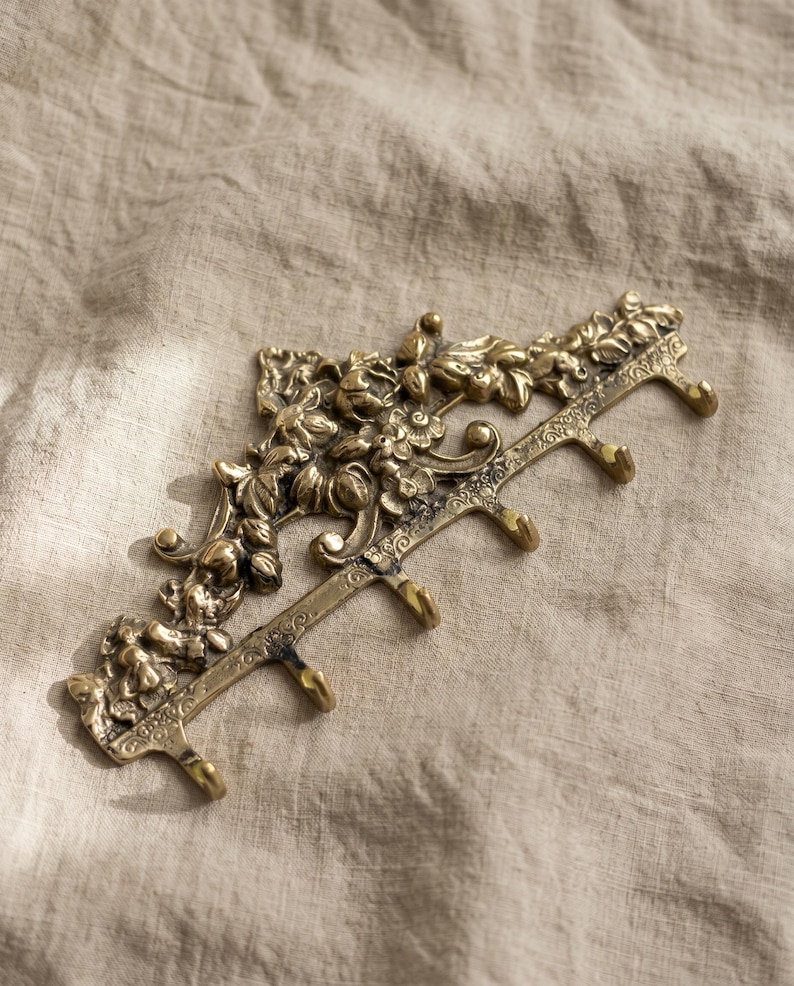 May include: A decorative brass wall hook with floral and leaf detailing. The hook has five individual hooks for hanging items. The brass has an aged appearance. The wall hook is designed to be mounted on a wall.