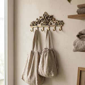 May include: A decorative brass wall hook with floral detailing, holding two beige linen bags. The hook has five curved hooks for hanging items. The bags are different sizes and have a natural, neutral color.